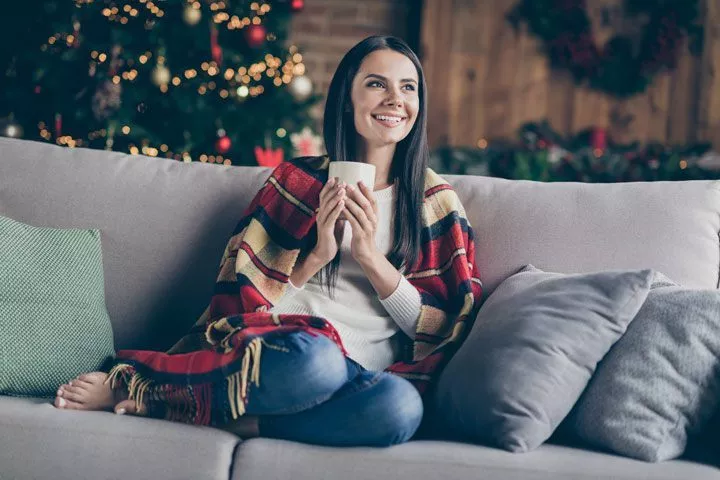pretty dark haired woman smiling while sitting on couch in room with Christmas tree - holiday season