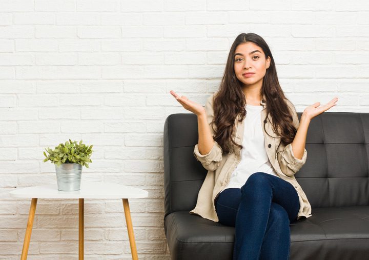 pretty brunette woman with long hair sitting on a couch holding her arms in a questioning gesture - resolutions or goals