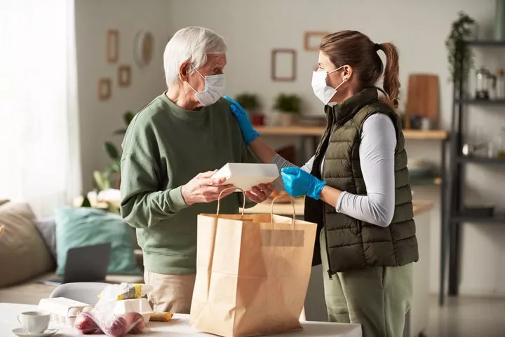 woman taking groceries to senior man - both donning masks - volunteering