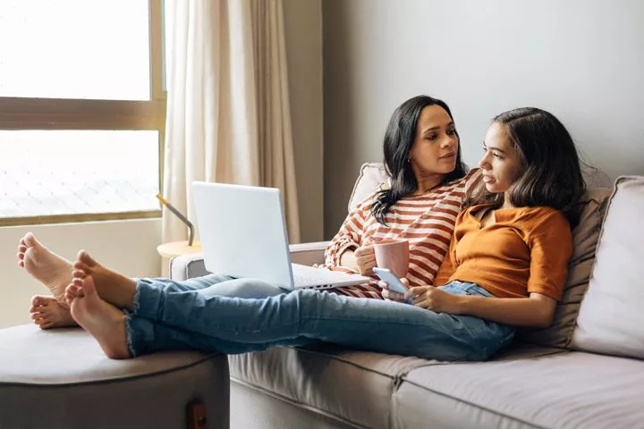 pretty Latinx mom sitting on couch at home with her preteen or young teenage daughter, while they look at a laptop together and talk casually - teens and mental health
