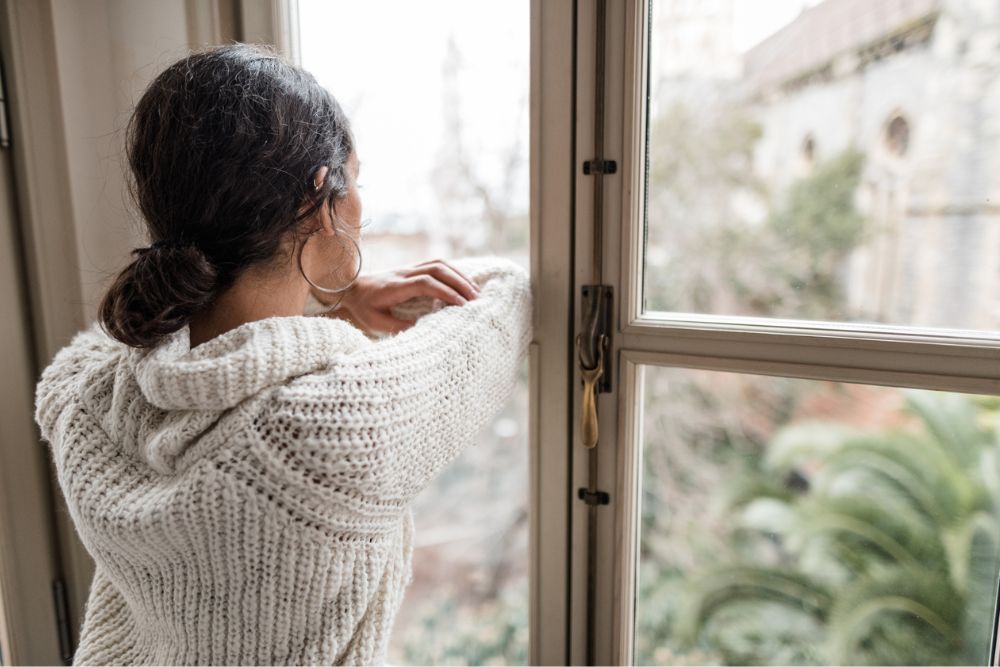 Woman in a cozy sweater looking out a window with a contemplative posture
