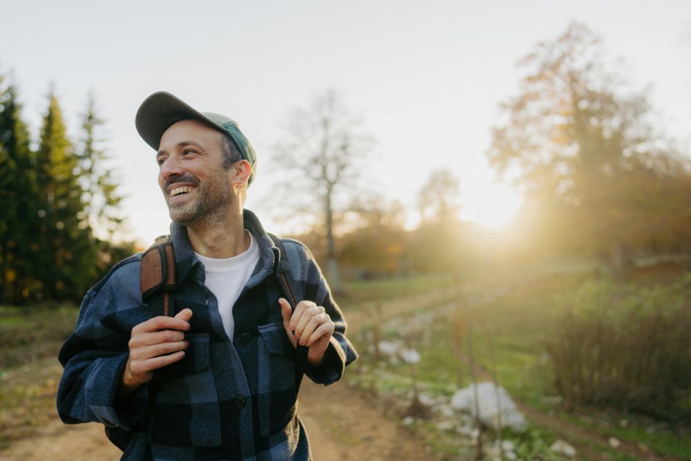 Smiling man wearing a cap and plaid jacket with a backpack, walking outdoors on a sunny day along a trail surrounded by trees and fields.