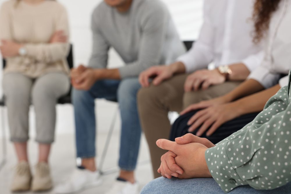 Close-up of a support group session, showing people seated in a circle with hands folded, suggesting a group therapy or recovery meeting setting