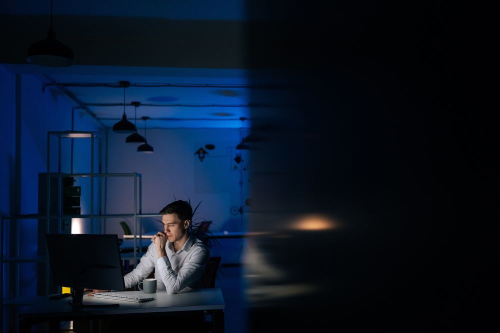 A man working late at a computer in a dark office, with only his desk light on, symbolizing overwork, hustle culture, or corporate burnout.