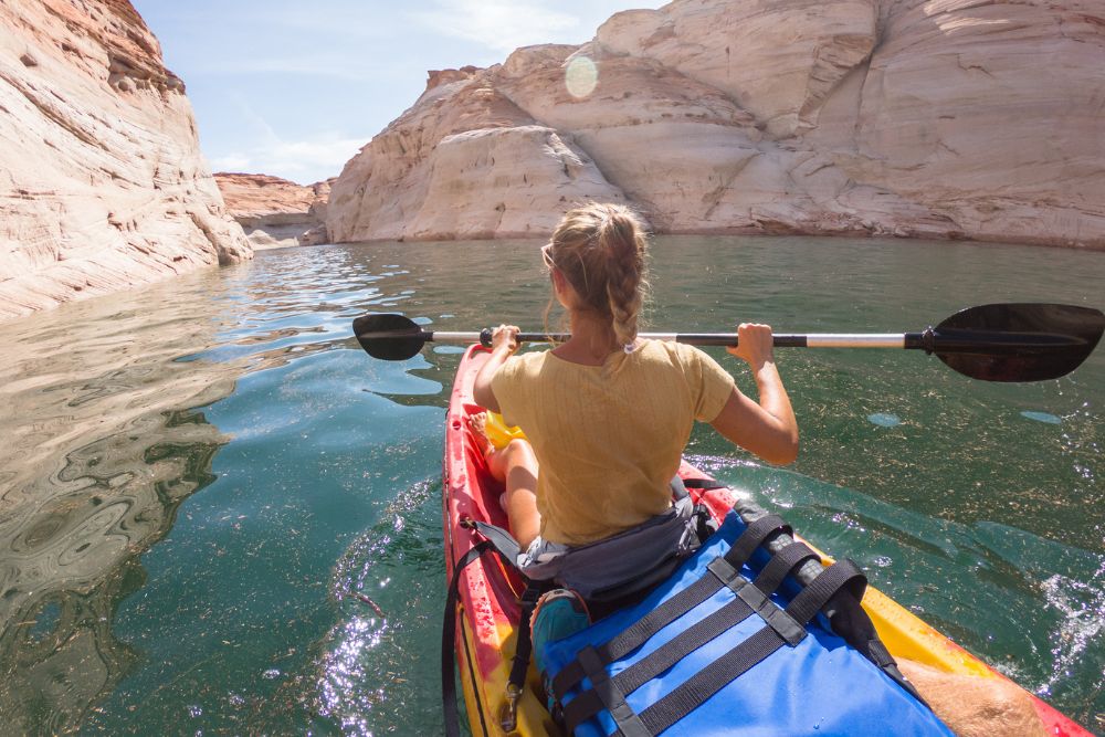 A woman paddles a red kayak through calm water surrounded by tall, sunlit rock formations. She faces forward, enjoying the peaceful summer adventure.