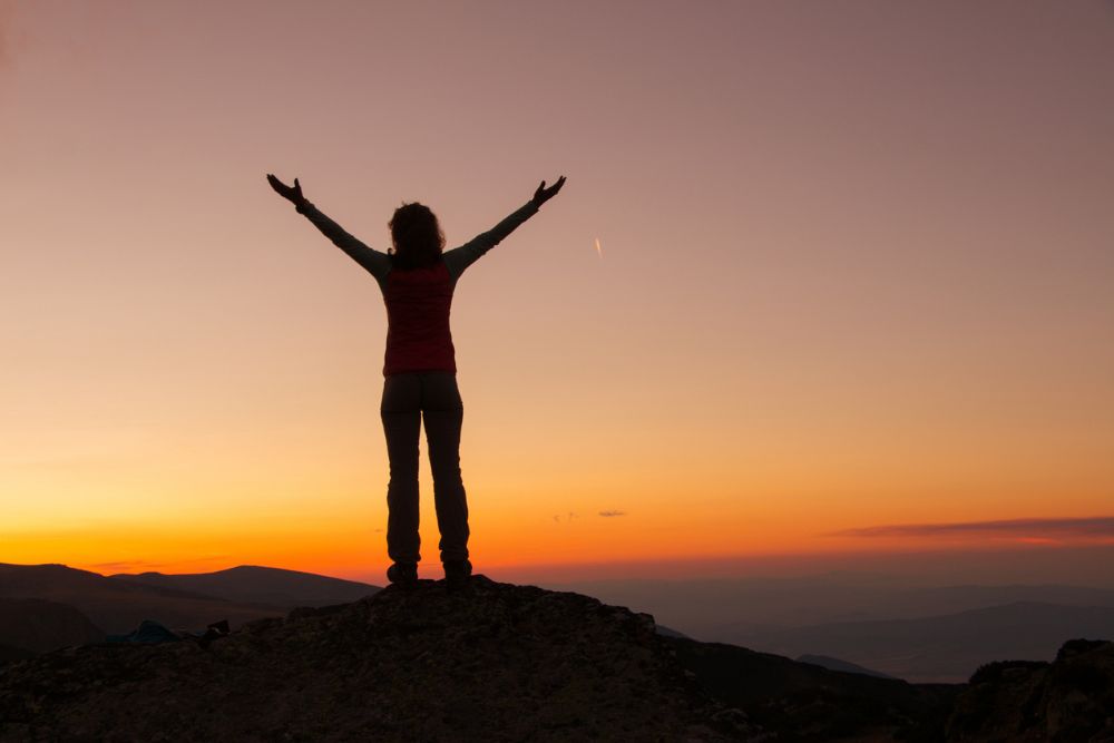 Silhouette of a person standing on a mountain with arms raised toward the sky at sunset, symbolizing hope, positivity, and empowerment.
