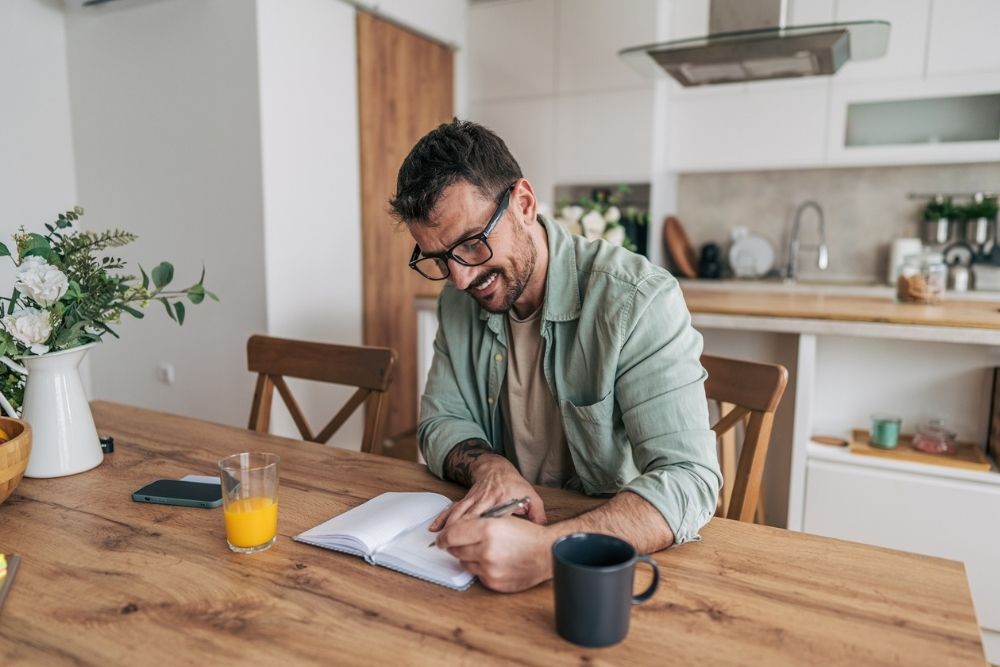 A man sits at a kitchen table smiling while writing in a journal, with a cup of coffee and a glass of juice nearby.