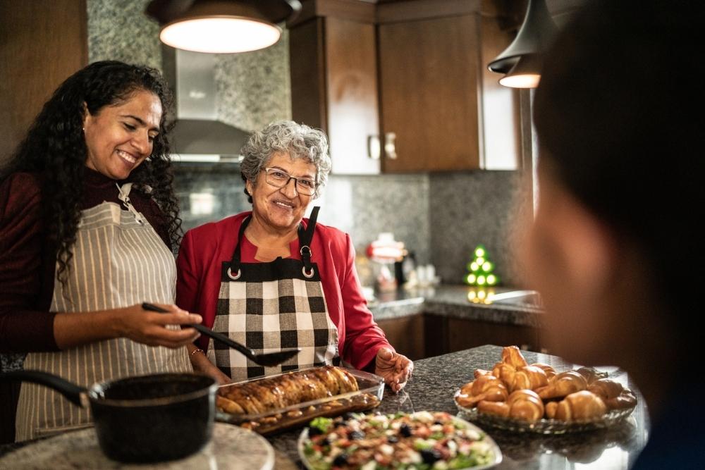Two women wearing aprons smile while preparing holiday food in a warm kitchen, surrounded by pastries, salad, and festive décor.