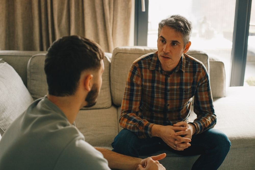 Two men sit on a couch engaged in a serious conversation, representing setting healthy boundaries, open communication, or counseling support.