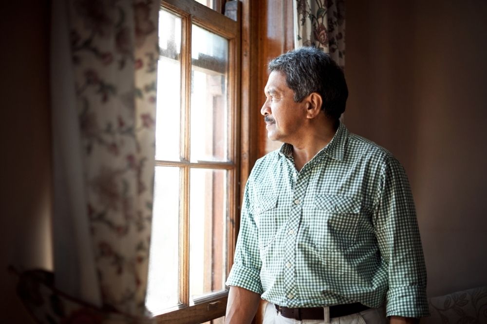 Man in a green checkered shirt stands by a window indoors, looking outside thoughtfully as sunlight filters in.